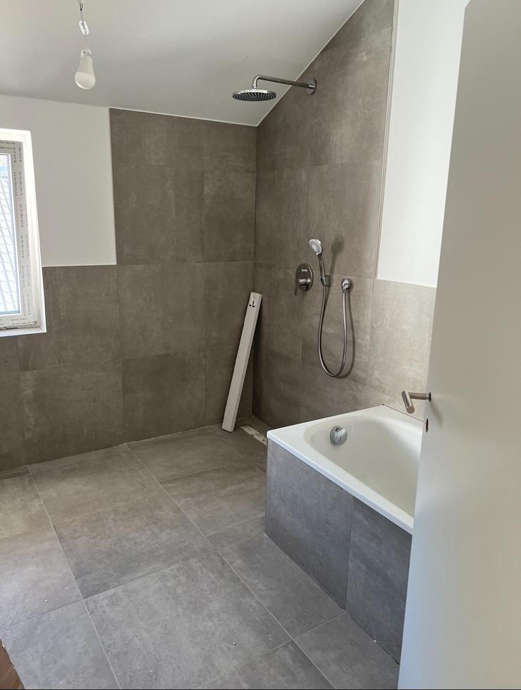 Frisch gefliestes Bad in Grau mit Wanne und Regendusche, Glühbirne hängt von der Decke