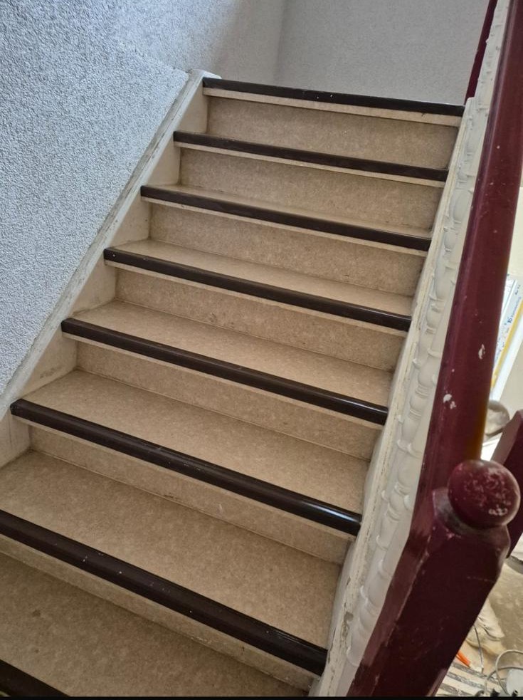 Alte Treppe mit abgenutztem beigem Belag und dunklen Kantenprofilen, roter Handlauf
